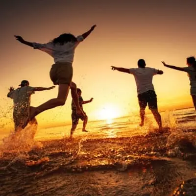 Five people run and splash in shallow water at the beach during sunset, silhouetted against the orange sky.