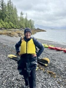 A person wearing a yellow life jacket and a beanie stands on a rocky shoreline by the water, with kayaks and paddles in the background.