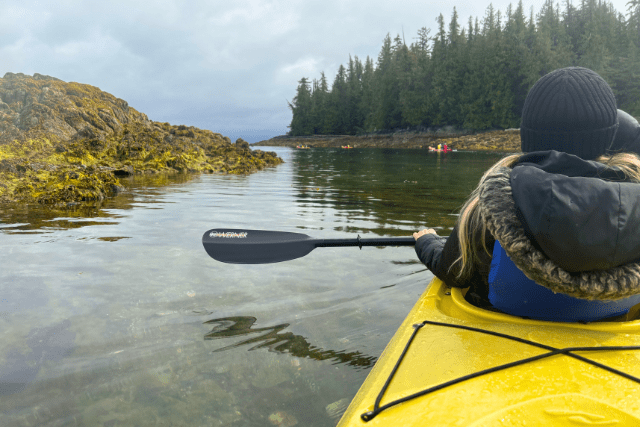Person paddling a yellow kayak in a calm coastal waterway, surrounded by rocky outcrops and evergreen trees on a cloudy day.