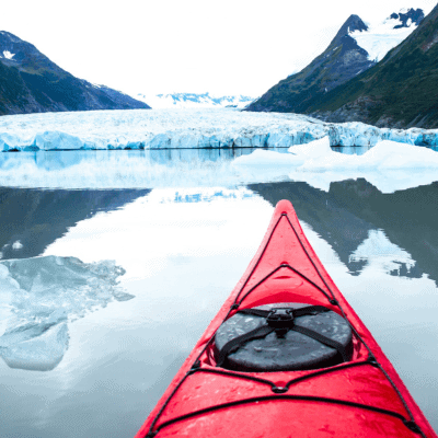 A red kayak on calm water approaches a glacier with icebergs floating nearby, surrounded by mountainous terrain.