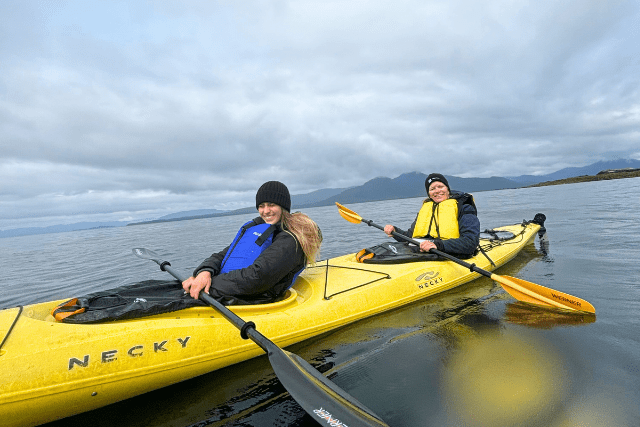Two people paddle a yellow tandem kayak on a calm body of water under a cloudy sky.