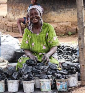 A person in a green dress sits outdoors, smiling, while handling pieces of charcoal placed in several buckets. A child is seated in the background.