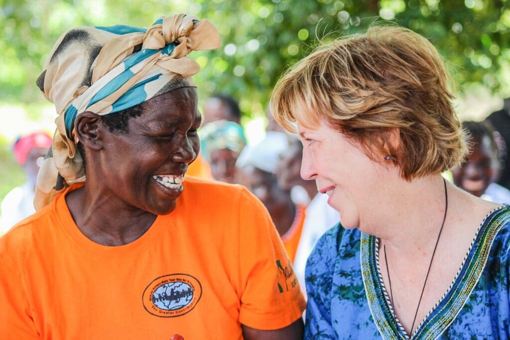 Two women smiling at each other outdoors, with one wearing an orange shirt and headscarf and the other in a blue top.