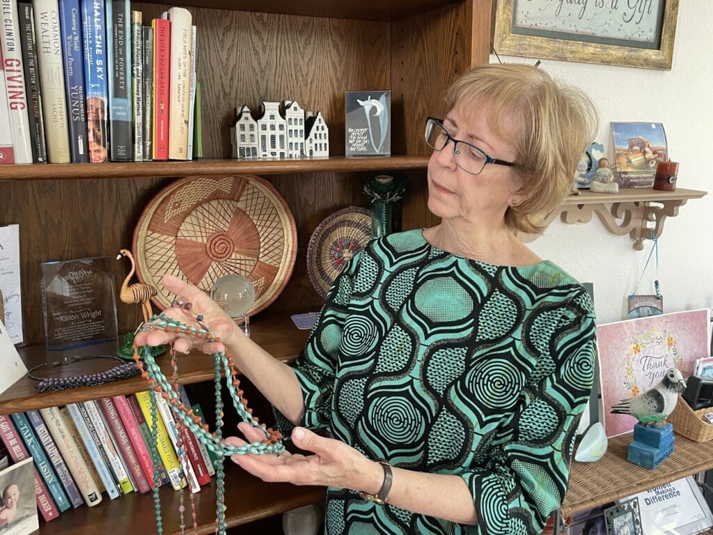A woman holding a beaded necklace stands in a room with a bookshelf filled with The Greater Contribution and decorative items.