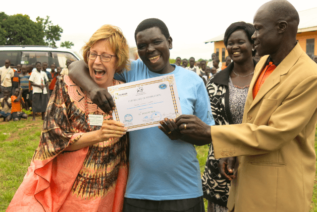 A group of people celebrate as one person holds a certificate. Another person laughs with an arm around them. There are onlookers and a vehicle in the background.