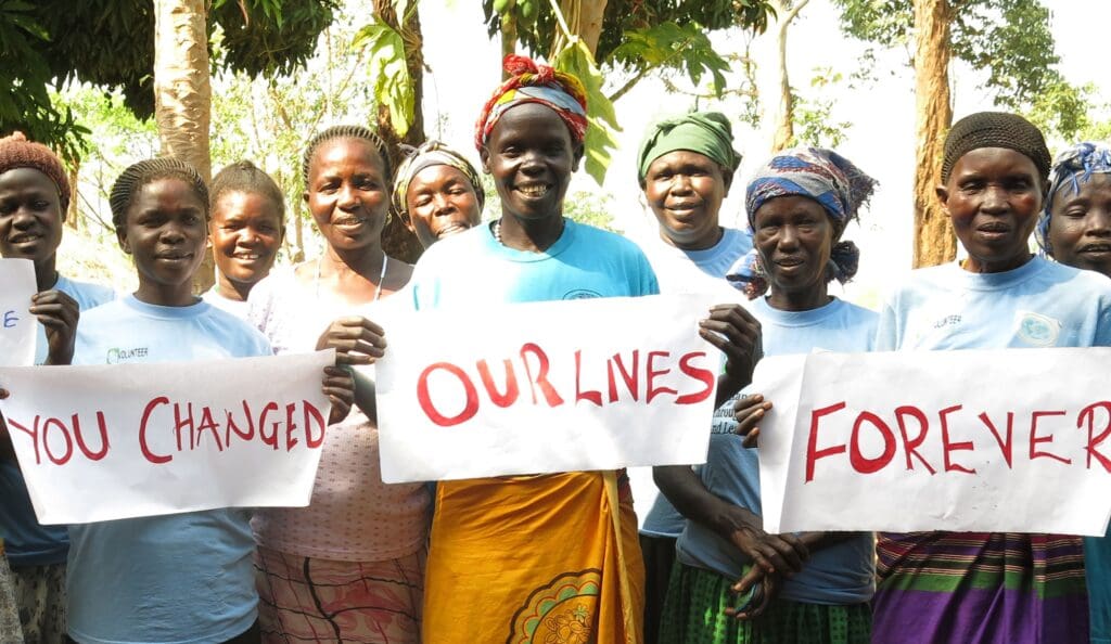 A group of smiling women holding signs that read "You changed our lives forever," standing outdoors with trees in the background.