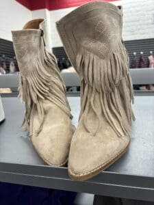 A pair of beige suede cowboy boots with fringe detailing, perfect for line dancing, displayed on a gray shelf.