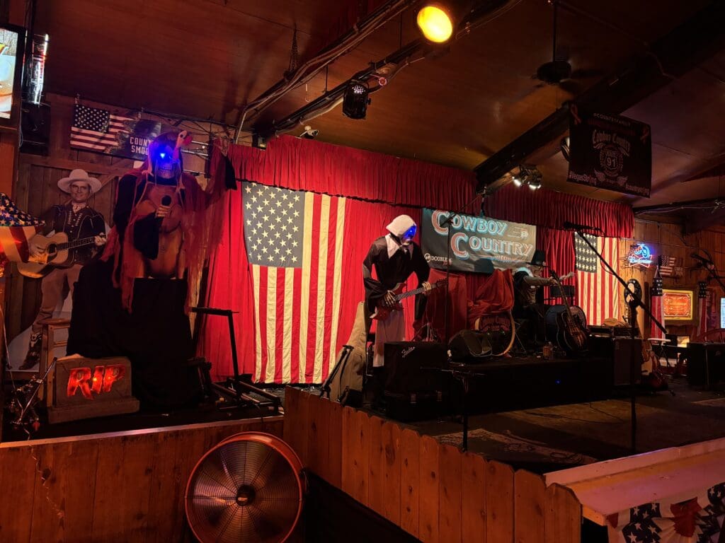 A dimly lit stage, adorned with Halloween decorations like skeletons and a coffin, sets the scene. An American flag hangs proudly in the background, while musical instruments await beside space for line dancing fun.