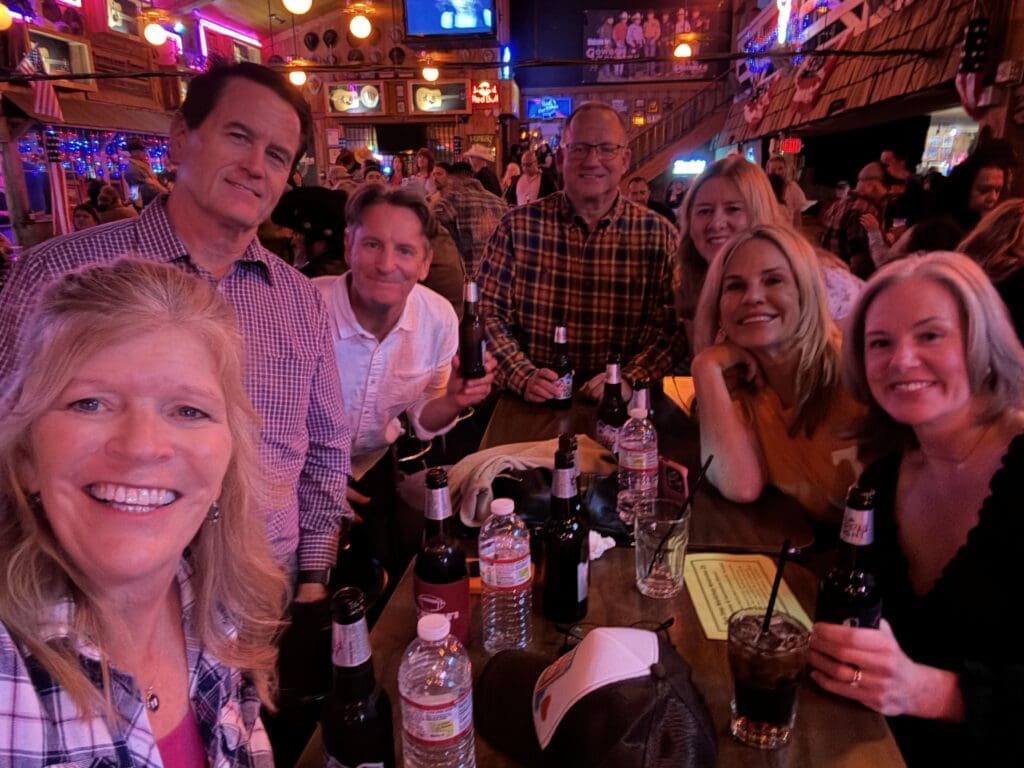 A group of people smiling at a table with drinks in a lively bar setting, the energetic rhythm of line dancing in the background adding to the joyful atmosphere.