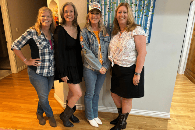 Four women standing indoors, smiling at the camera, wearing casual outfits and boots perfect for line dancing.