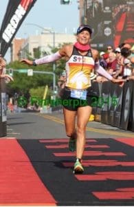 A triathlete smiles with arms outstretched as she crosses the finish line at an Ironman race, surrounded by cheering spectators.