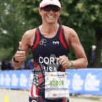 A triathlete wearing a USA uniform and sunglasses runs outdoors, smiling and giving two thumbs up, with trees and event barriers in the background.