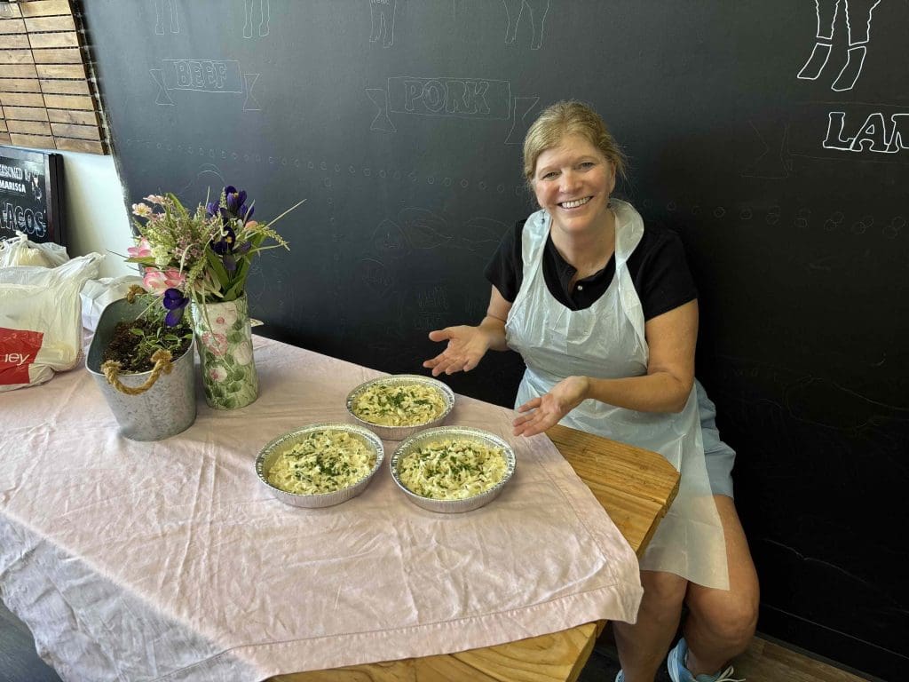 A woman wearing an apron sits at a table displaying three uncooked quiches. There are flowers in a vase and a chalkboard wall in the background.