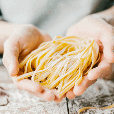 Hands holding a bundle of uncooked fettuccine pasta with flour dusted on the noodles and wooden surface.