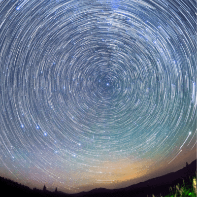 Long-exposure photograph of the night sky showing circular star trails above a dark, mountainous landscape with a faint orange horizon.