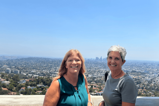 Two women stand in front of a cityscape on a clear day, with a distant skyline and buildings visible under a blue sky.