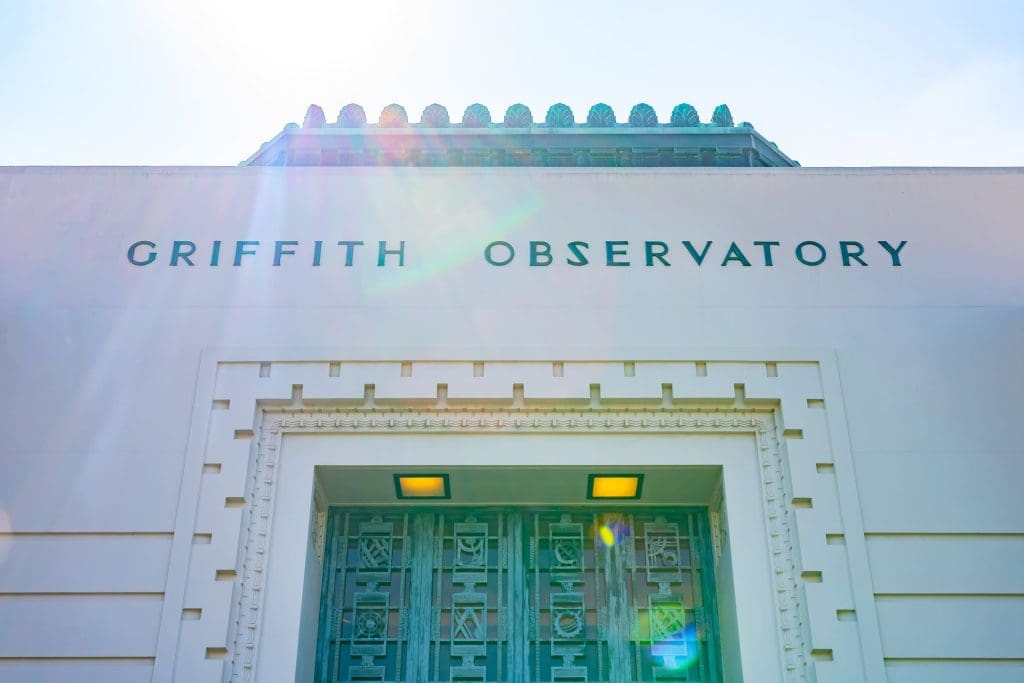 The exterior of Griffith Observatory is shown from a low angle, with sunlight creating a lens flare across the building’s name above the entrance.