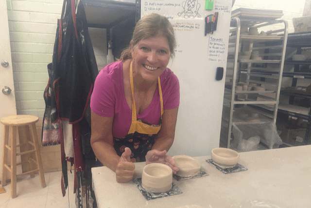 A woman in a pottery studio smiles at the camera while shaping clay bowls on a table. Shelves with pottery and supplies are visible in the background.