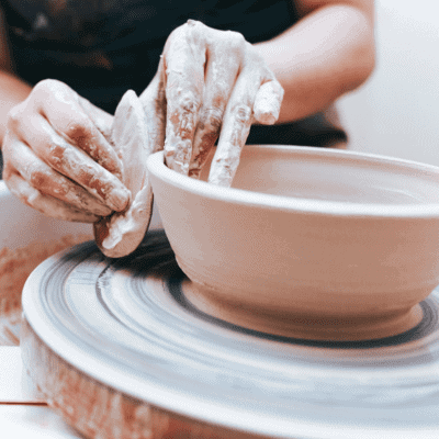 Close-up of hands shaping a clay bowl on a pottery wheel, with one hand smoothing the edge using a tool.